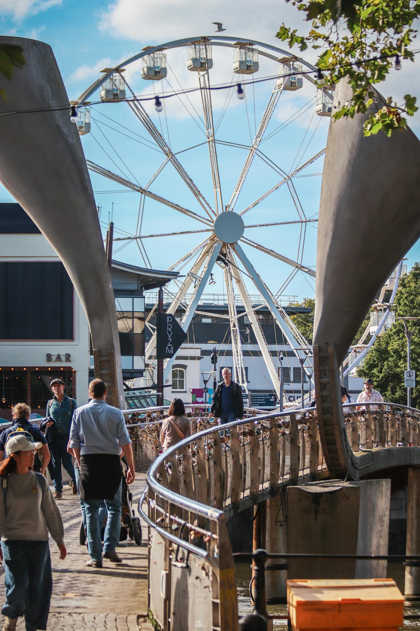 Crowd enjoying a sunny day at Bristol Harbourside with a Ferris wheel in view.